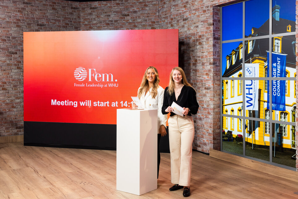 Two smiling smartly-dressed young women with long blonde hair stand behind a white podium in front of a large red sign which says "Fem. Female Leadership at WHU"