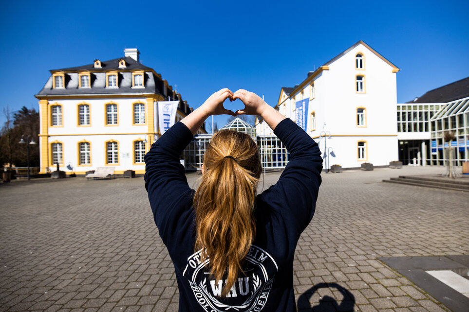 A young women wearing a blue WHU seatshirt and with her hair in a ponytail sits with her back to the camera on the Burgplatz at WHU Campus Vallendar. She holds up her arms and makes the shape of a heart with her hands.