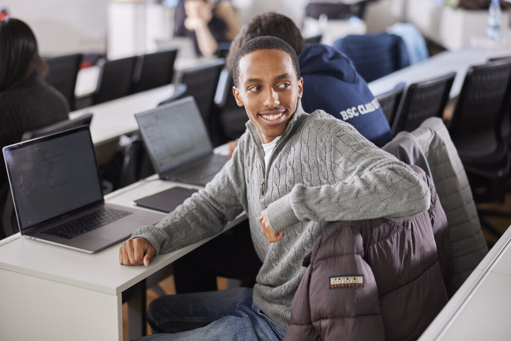 A young student in a gray sweater sits in front of his notebook in the lecture hall. He rests his hand on the back of his chair and turns to the person behind him with a broad smile on this face.