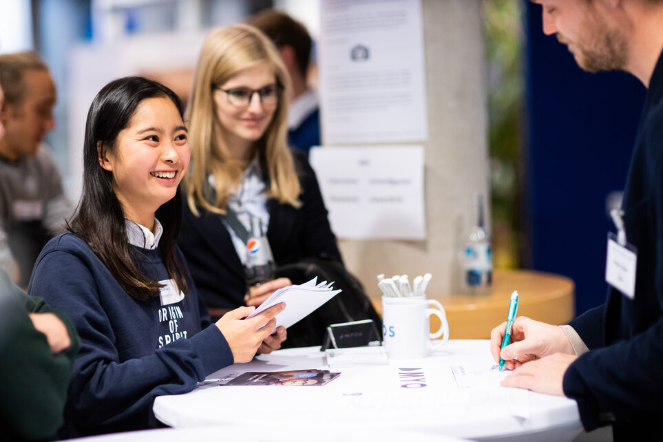 A smiling young woman with long dark hair stands at a table at Founders Career Day and receives information from a man who is writing something.