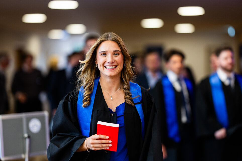 A woman in a graduation gown proudly smiling and holding a red book.