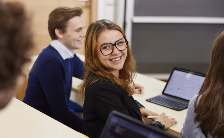 A woman with glasses happily smiles while sitting in front of a laptop.
