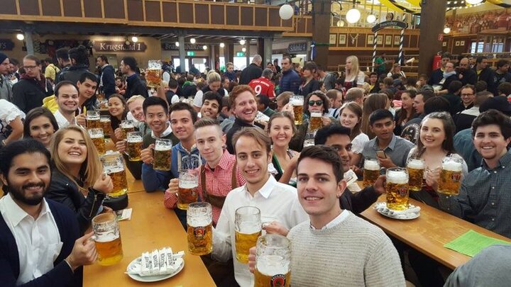 Viele lachende junge Leute mit Bierkrug im Hand sitzen im Bierzelt beim Oktoberfest in München