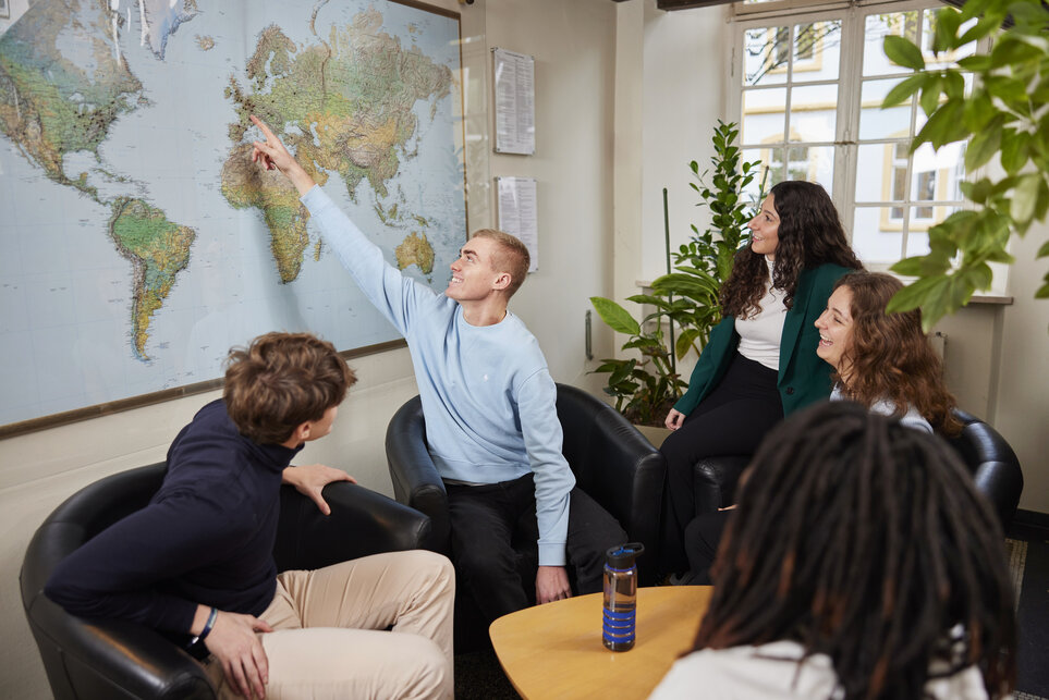 Ein junger Mann im blauen Hemd steht vor einer Weltkarte an der Wand und zeigt auf Deutschland während vier weitere Studierende schauen zu