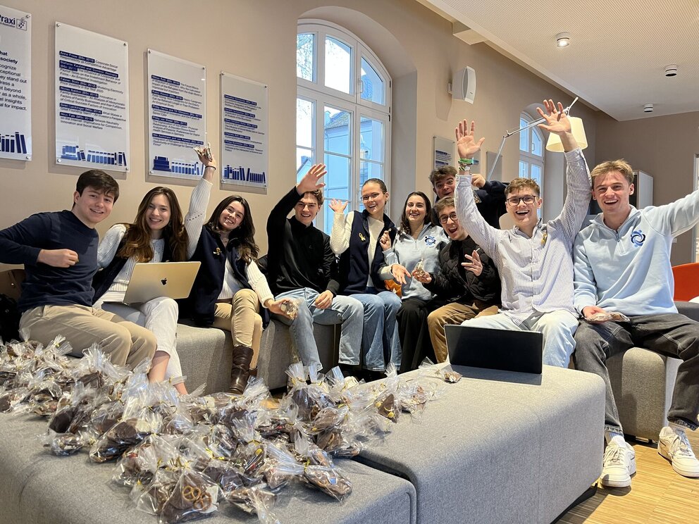 A group of ten young people sit on grey sofas facing the camera. They are laughing and some raise their arms. There is a  pile of transparent gift bags in front of them 