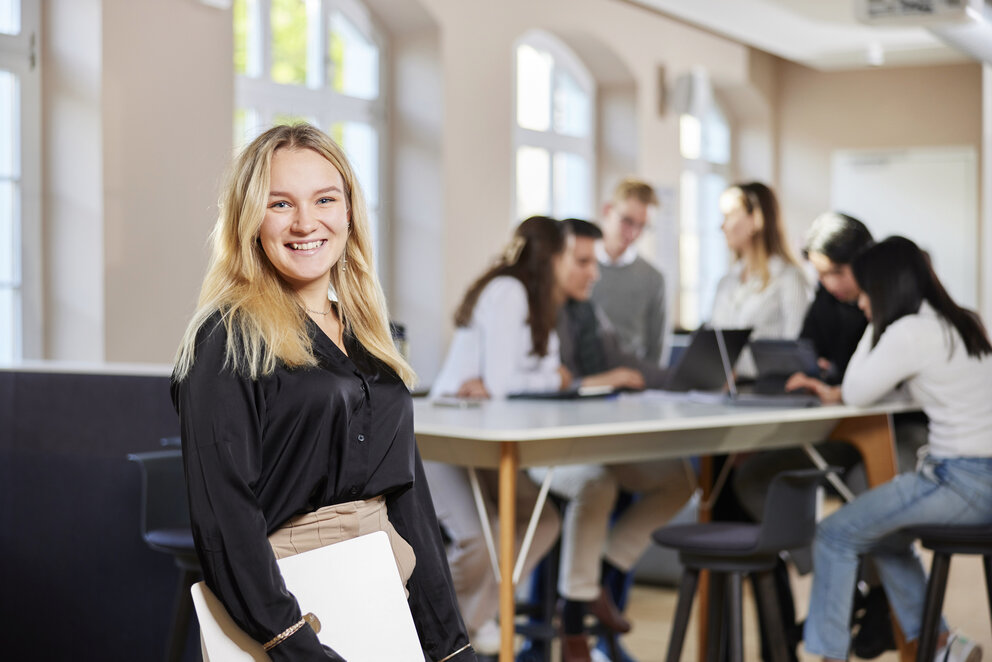 Eine junge Frau mit langen blonden Haaren, die eine langärmelige schwarze Bluse trägt und einen weißen Ordner in der Hand hält, steht lächelnd vor der Kamera. Im Hintergrund sitzen sechs Studierende an einem Tisch und arbeiten an zwei aufgeschlagenen Notebooks.