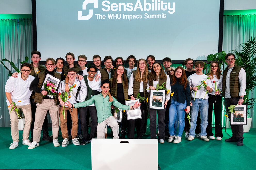 Eine Gruppe von etwa 20 jungen Menschen posiert und lächelt für die Kamera vor einem großen Bildschirm, auf dem SensAbility - The WHU Impact Summit steht. Viele von ihnen halten bunte Blumen in der Hand.